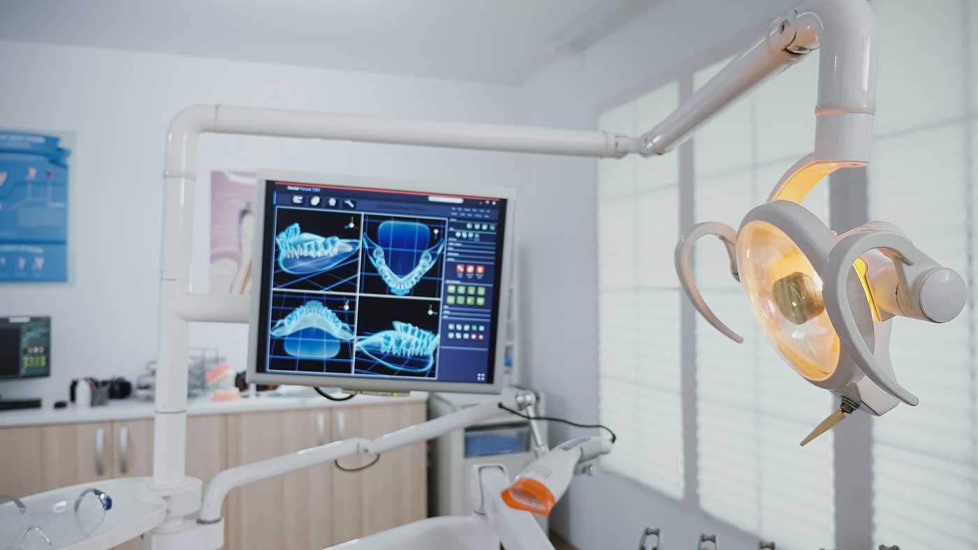 Ein Behandlungszimmer in einer Zahnarztpraxis mit moderner Ausstattung. Im Vordergrund ist eine beleuchtete OP-Lampe zu sehen, daneben ein Monitor, auf dem digitale Röntgenbilder von Zähnen und Kieferknochen angezeigt werden. Im Hintergrund befinden sich Schränke und medizinische Geräte. Der Raum ist hell und aufgeräumt.