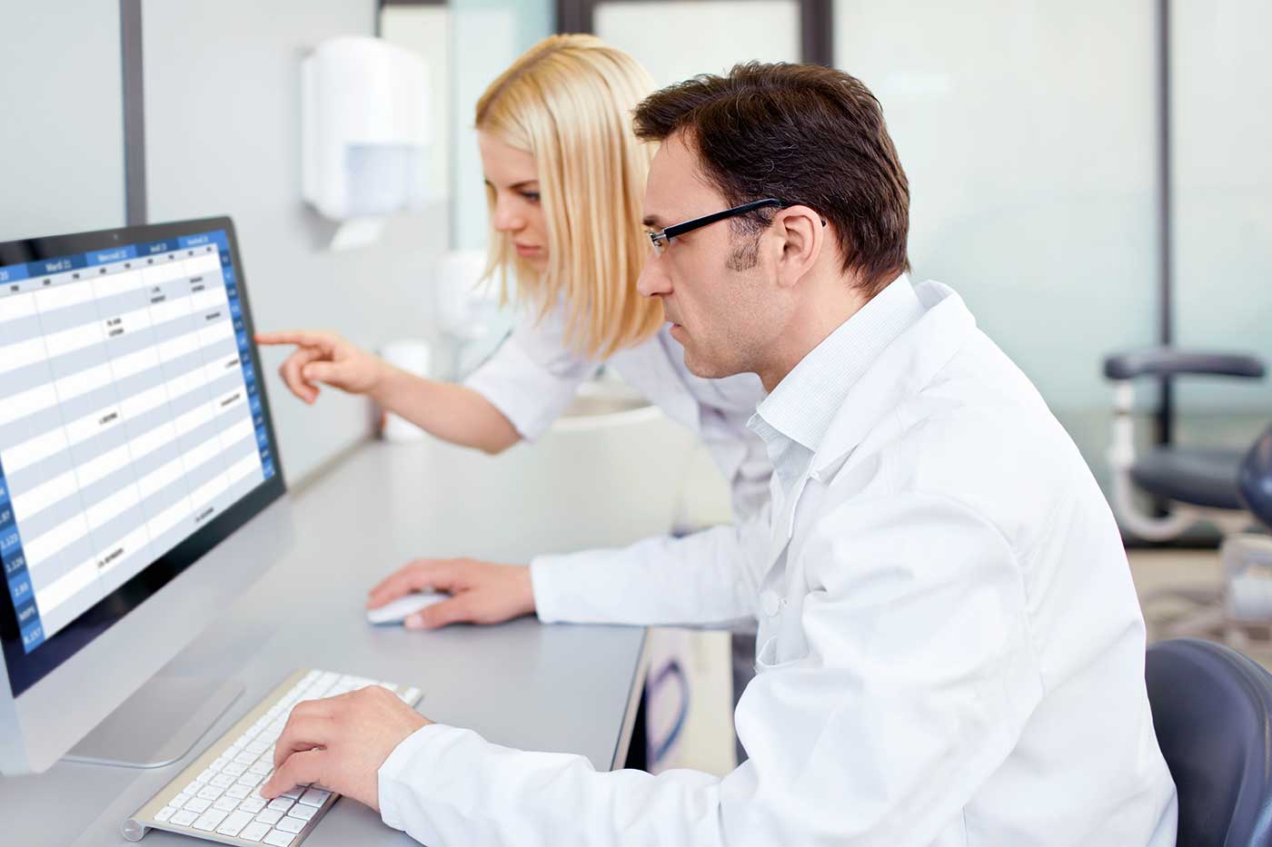 Zwei Personen in weißen Kitteln arbeiten an einem Computerarbeitsplatz in einer medizinischen Einrichtung. Eine Frau zeigt auf den Bildschirm, während ein Mann die Tastatur und Maus bedient. Auf dem Monitor ist eine Tabelle mit Terminen oder Patientendaten zu sehen. Im Hintergrund sind weitere Einrichtungsgegenstände einer Praxis sichtbar.