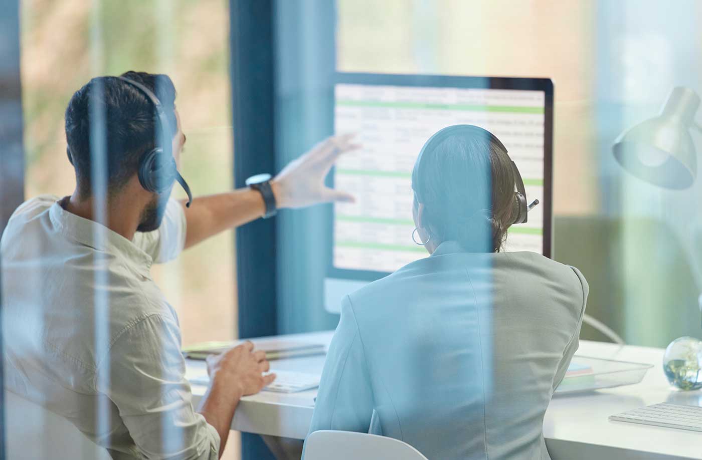 Zwei Personen mit Headsets sitzen an einem Schreibtisch vor einem großen Monitor. Eine Person zeigt auf den Bildschirm, auf dem eine Tabelle mit grün-weißen Einträgen zu sehen ist. Die Szene wird durch Glaswände aufgenommen, wodurch Spiegelungen sichtbar sind. Auf dem Schreibtisch liegen Dokumente, eine Tastatur und eine kleine Weltkugel.