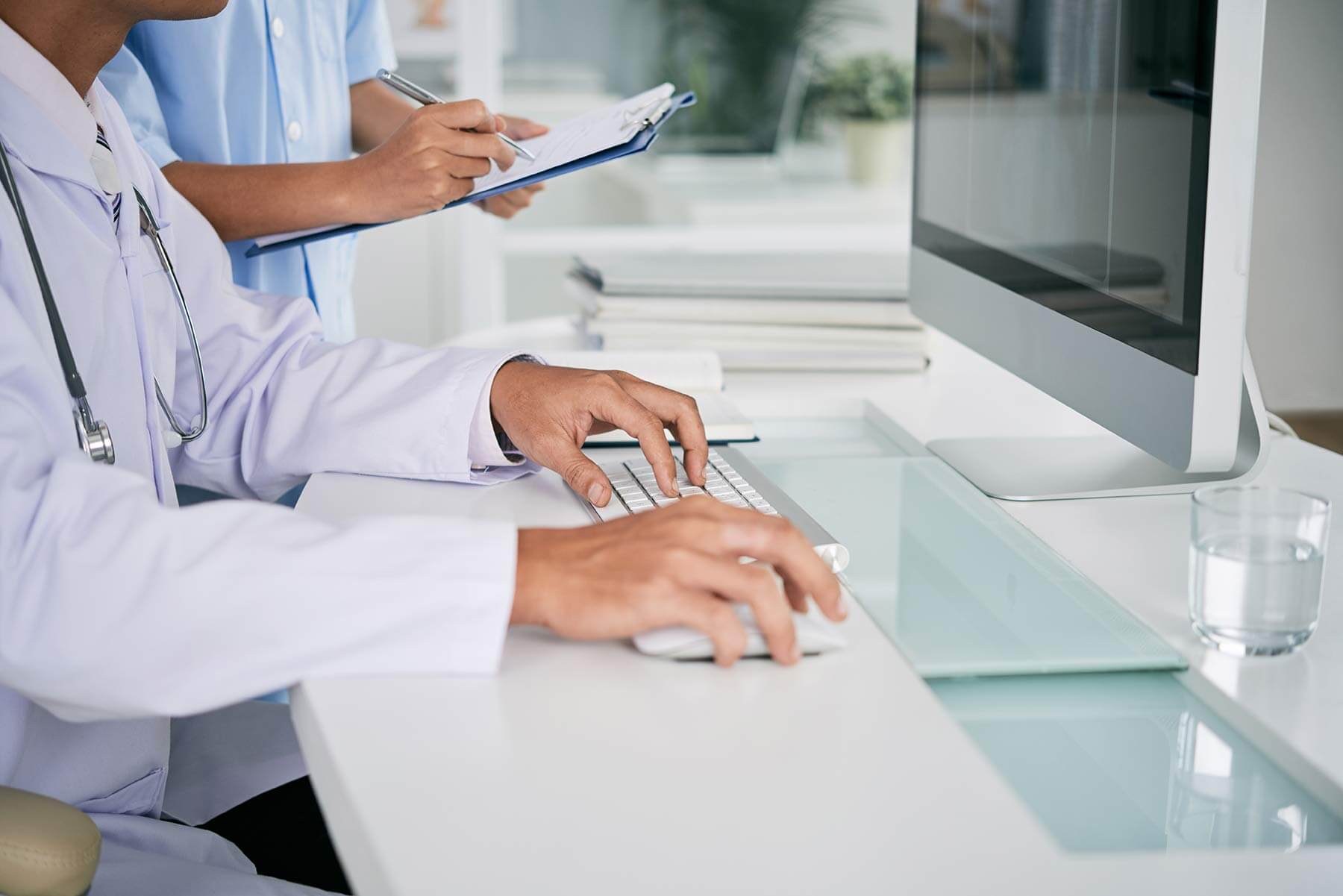 Zwei Personen in medizinischer Kleidung an einem Schreibtisch mit Computer. Eine Person tippt auf einer Tastatur, während die andere auf einem Klemmbrett Notizen macht. Auf dem Tisch liegen Bücher und ein Glas Wasser, im Hintergrund ist eine Pflanze zu sehen.