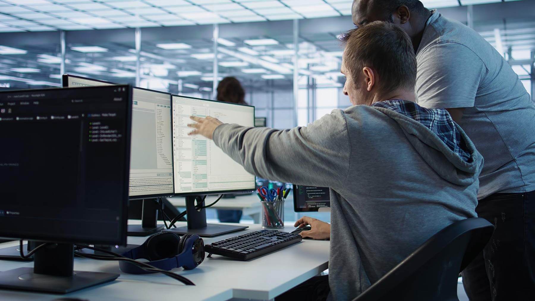 Zwei Personen arbeiten in einem modernen Büro an mehreren Monitoren. Eine Person sitzt am Schreibtisch, zeigt mit der Hand auf einen der Bildschirme, während die andere Person daneben steht und zuschaut. Auf den Monitoren sind Programmier- und Datenbankoberflächen zu sehen. Auf dem Schreibtisch befinden sich eine Tastatur, eine Maus, ein Kopfhörer und ein Behälter mit Stiften. Im Hintergrund ist ein heller, offen gestalteter Büroraum mit Glaswänden erkennbar.