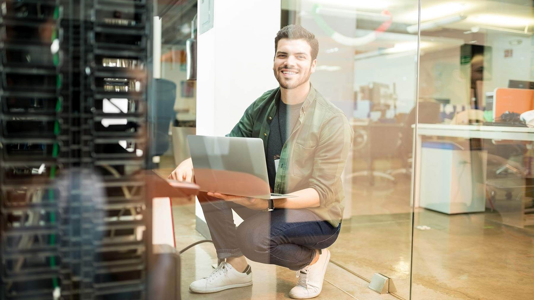 Ein junger Mann hockt lächelnd mit einem aufgeklappten Laptop in der Hand neben einem Serverrack in einem modernen Büro. Er trägt Freizeitkleidung und sitzt hinter einer Glaswand. Im Hintergrund sind Schreibtische, Bürostühle und technische Geräte sichtbar. Das Büro wirkt offen und hell.
