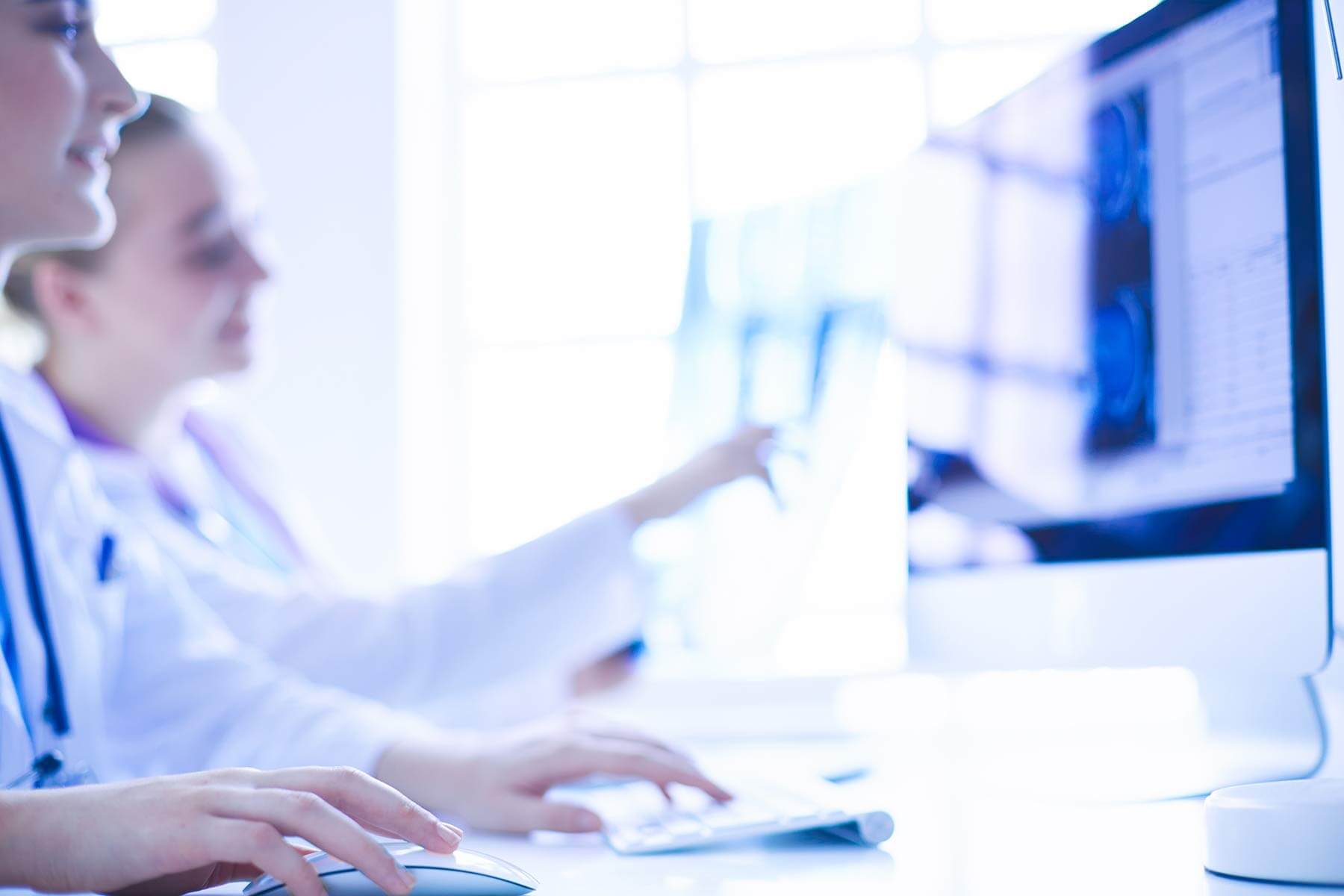 Zwei medizinische Fachkräfte in weißen Kitteln arbeiten an Computern. Eine Person bedient Tastatur und Maus, während die andere auf einen Bildschirm mit medizinischen Bilddaten zeigt. Die Umgebung ist hell und modern, mit Tageslicht, das durch große Fenster einfällt.