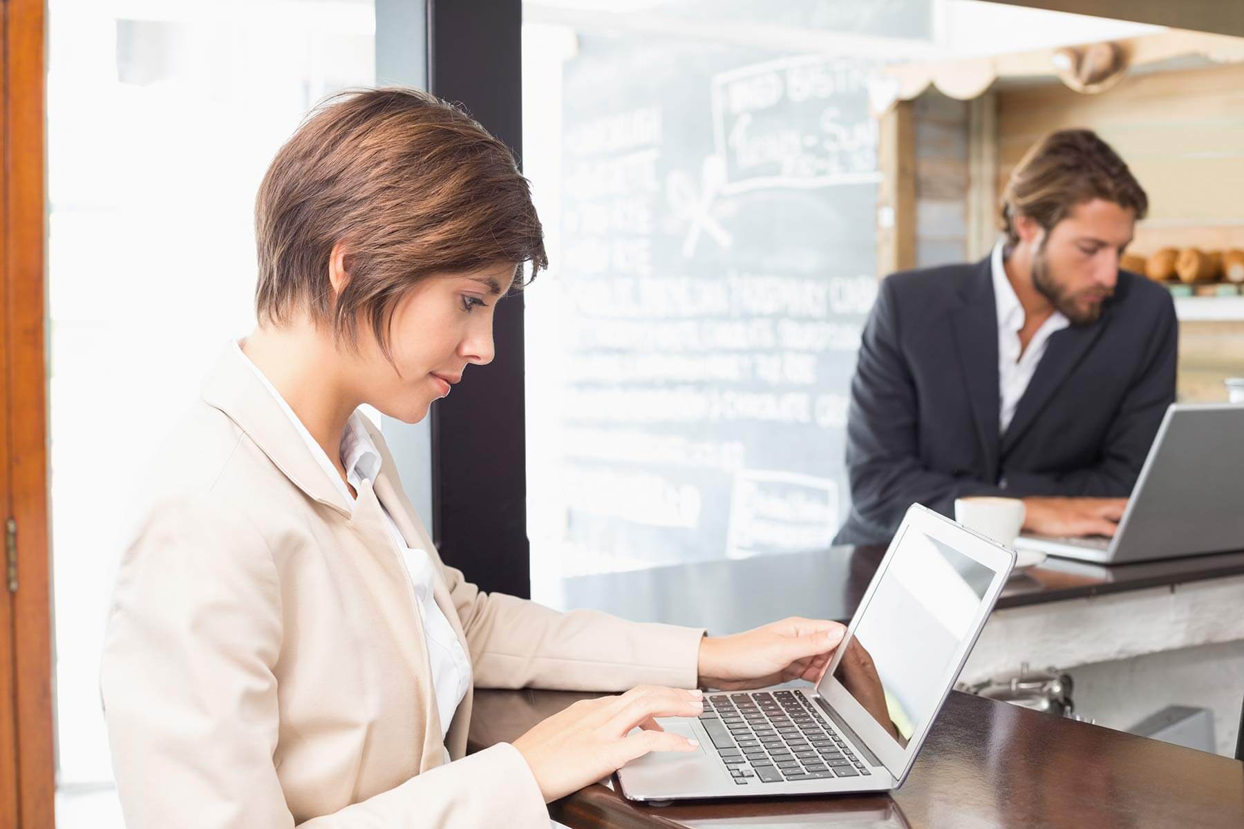 Zwei Personen in Businesskleidung arbeiten an Laptops in einem modernen Café oder Coworking-Space. Eine Person sitzt im Vordergrund, die andere im Hintergrund an einem separaten Tisch.