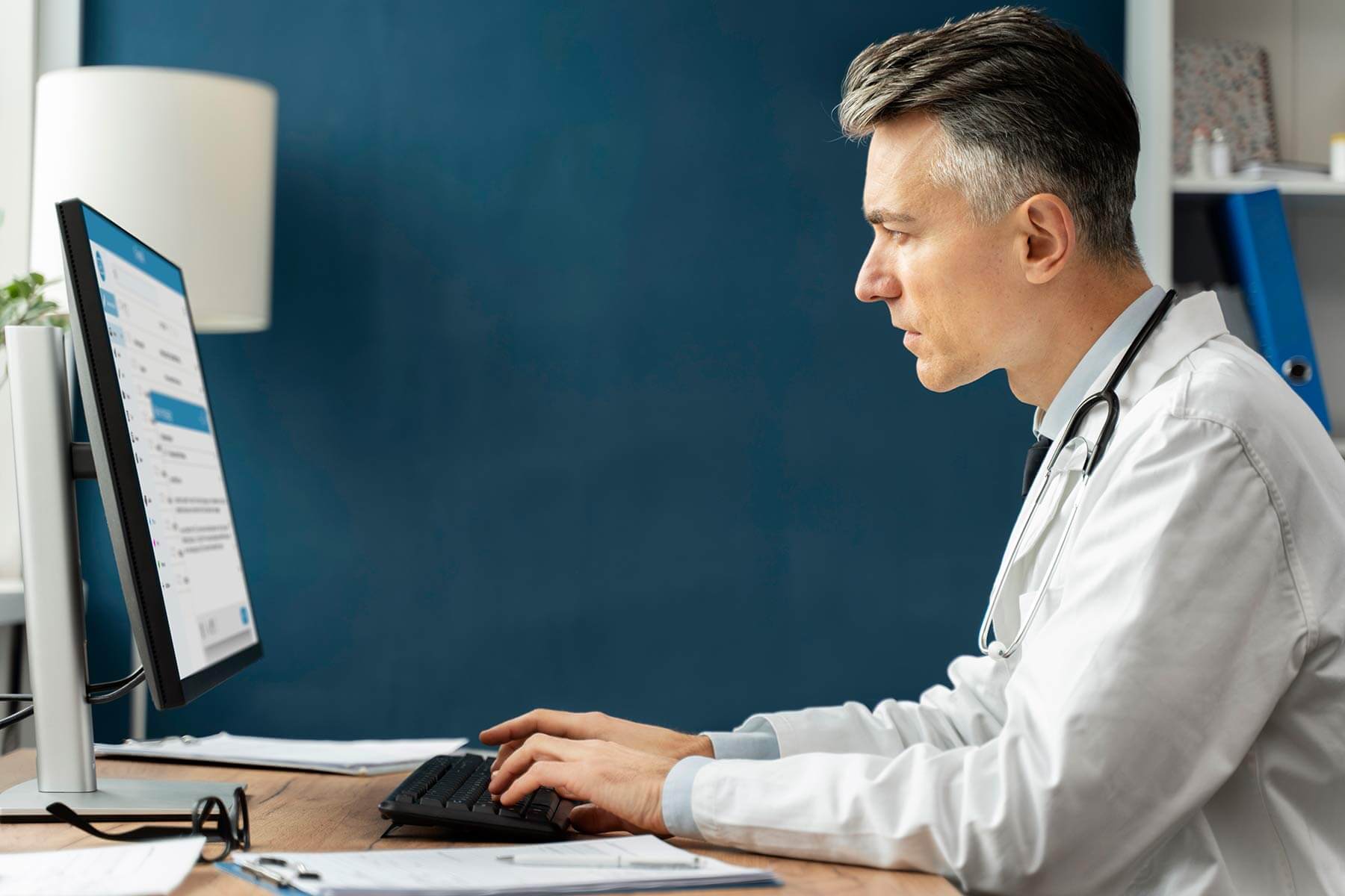 Ein Mann in weißem Kittel mit Stethoskop um den Hals sitzt an einem Schreibtisch und arbeitet am Computer. Auf dem Tisch liegen Dokumente, eine Brille und ein Stift.
